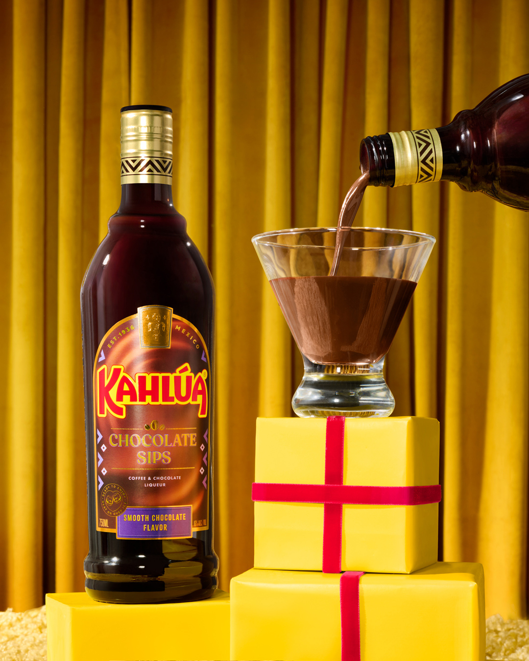 Kahlua chocolate sips liqueur being poured into a martini glass on yellow wrapped gifts in front of a gold curtain.