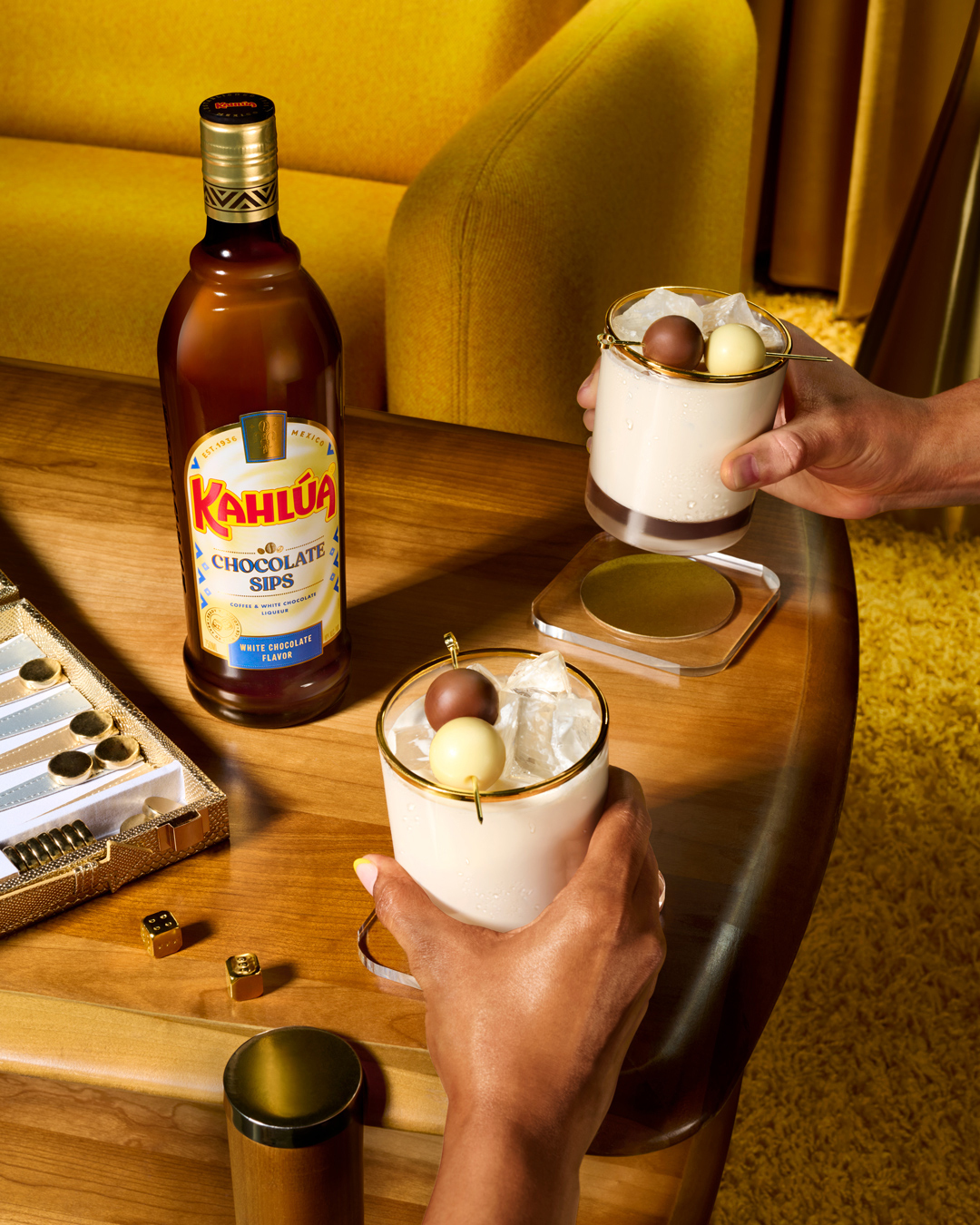 Kahlua Chocolate Sips liqueur bottle and two cocktails with chocolate balls, coasters, and game on a wood table with yellow chair.