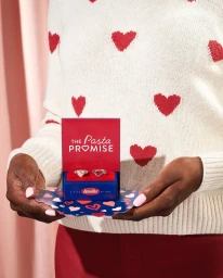 Person wearing a white sweater with red hearts holding open a small red promotional ring box reading The Pasta Promise.