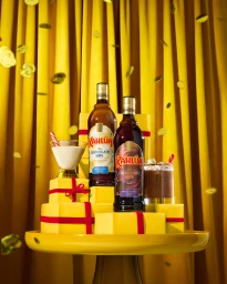 Two Kahlua Chocolate Sips bottles on yellow gift boxes with cocktails and floating coffee beans against yellow curtains