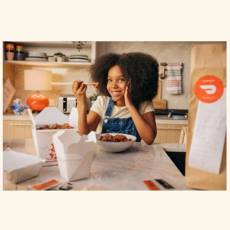 Young African-American girl enjoys a meal of Chinese takeout at home, using chopsticks and wearing denim overalls. Natural lighting.