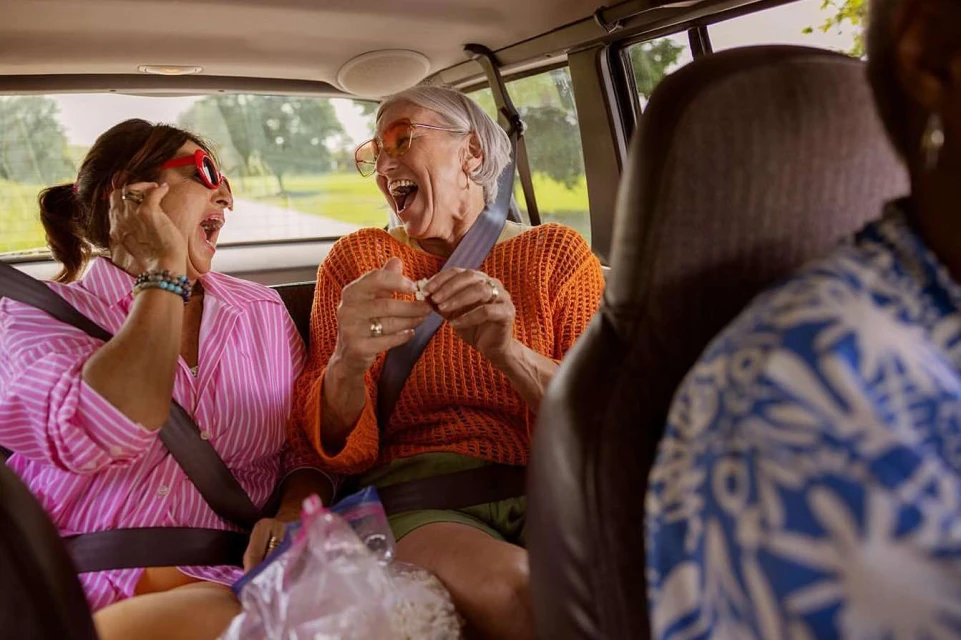 Two older women with glasses laugh in a car, one with pink striped shirt, the other in orange, road visible in the background.