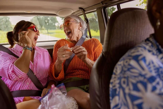 Two older women with glasses laugh in a car, one with pink striped shirt, the other in orange, road visible in the background.