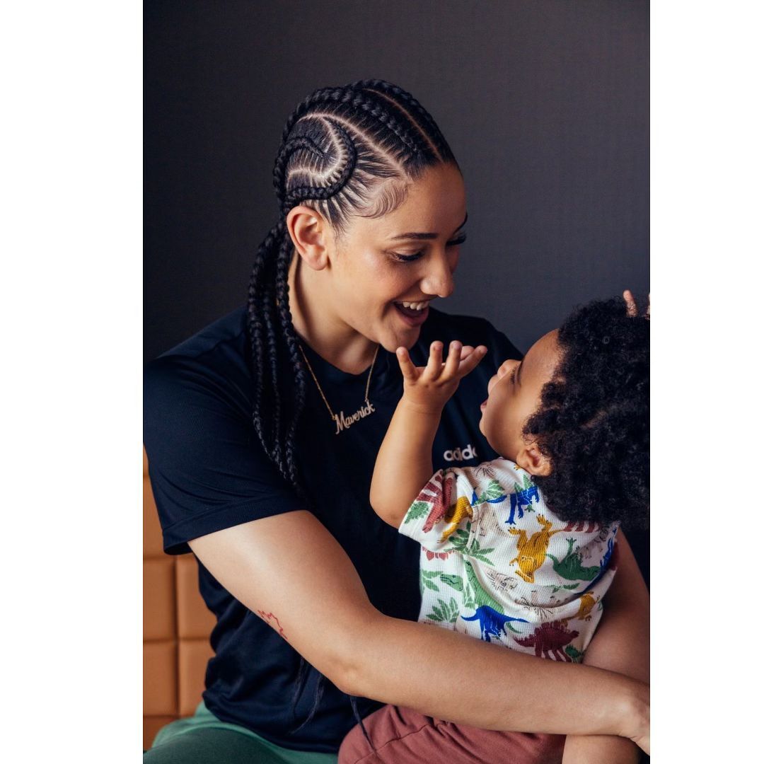 A smiling woman with braided hair holds a child with curly hair indoors against a dark gray backdrop, creating a warm and engaging portrait.