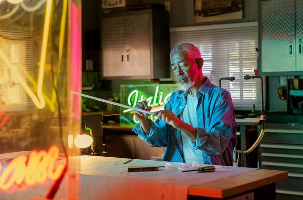 A mature man with greying hair is crafting a neon sign in his workshop, wearing a denim shirt. He is illuminated by colorful neon lights.