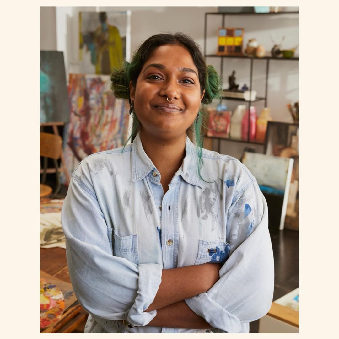 Smiling artist in a paint-splattered shirt stands in an art studio with paintings and art supplies. She has pigtails and a nose ring.