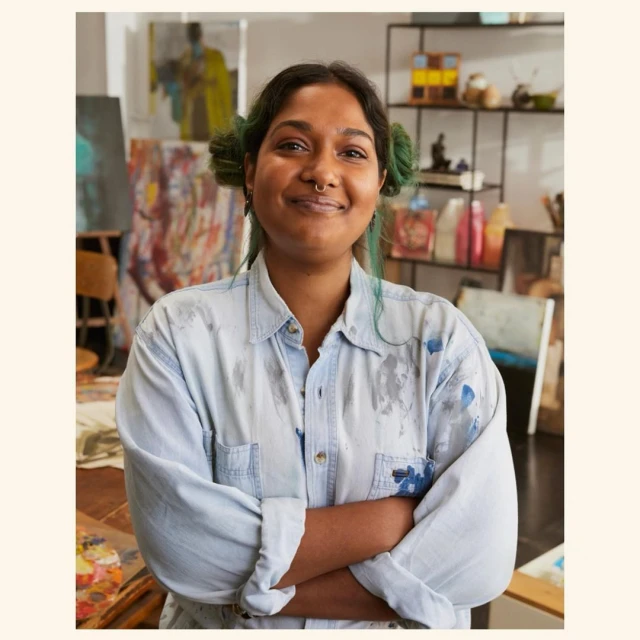 Smiling artist in a paint-splattered shirt stands in an art studio with paintings and art supplies. She has pigtails and a nose ring.