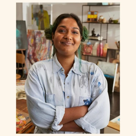 Smiling artist in a paint-splattered shirt stands in an art studio with paintings and art supplies. She has pigtails and a nose ring.
