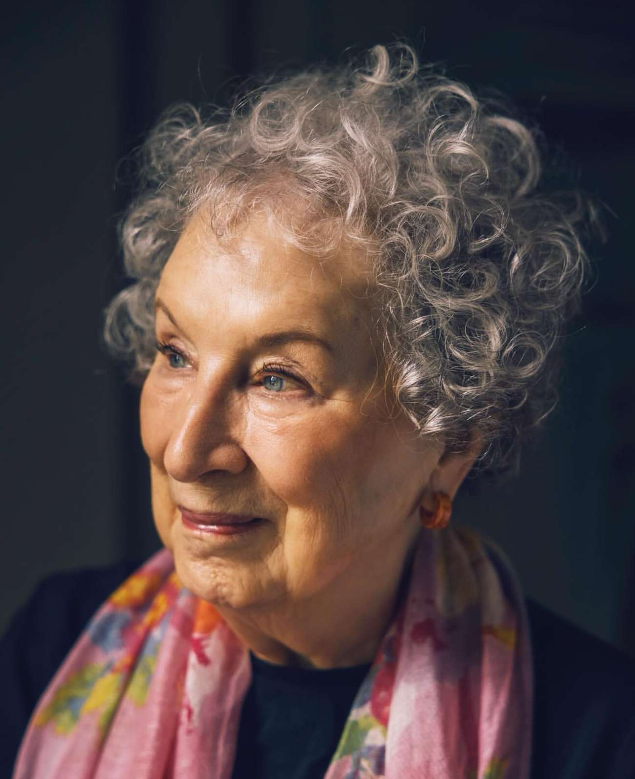 Portrait of an elderly woman with curly grey hair wearing a floral scarf and earrings looking off to the side with a slight smile.