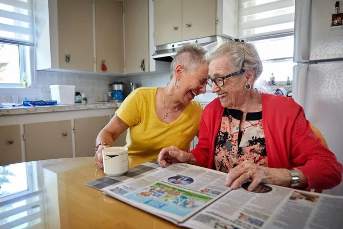 Two cheerful elderly women share a laugh while reading a newspaper at a sunlit kitchen table, creating a warm and joyful scene.