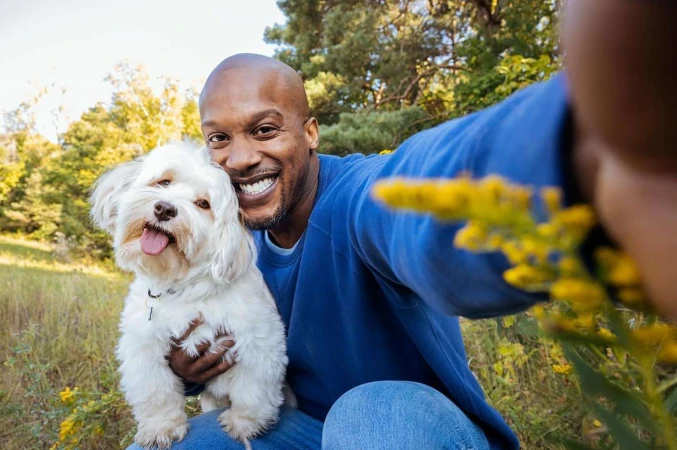 A smiling black man wearing a blue sweatshirt holding a fluffy white dog while taking a selfie outdoors.