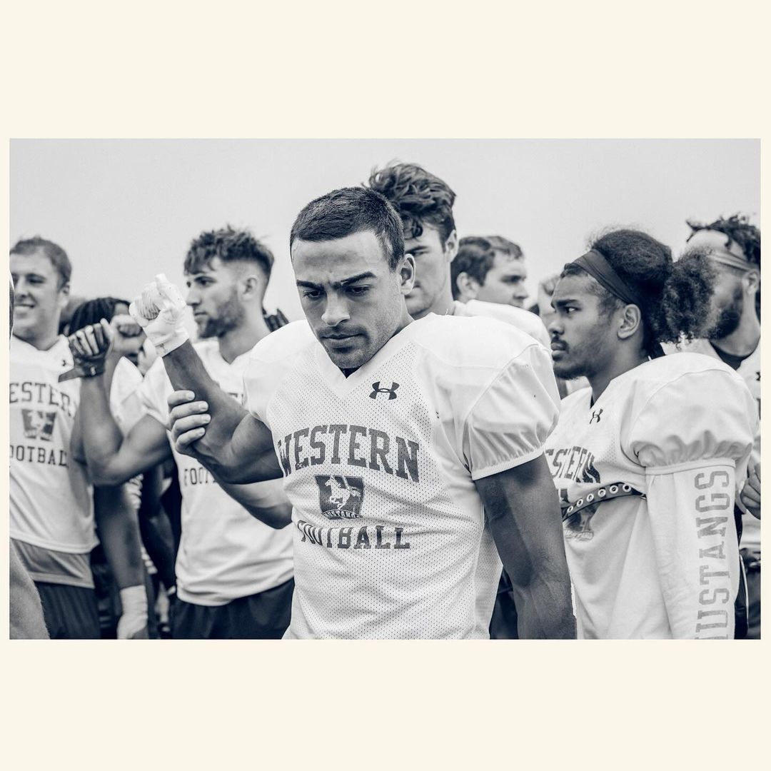 Black and white shot of Western Mustangs football players in uniform, focus on athlete in the foreground, sky background.