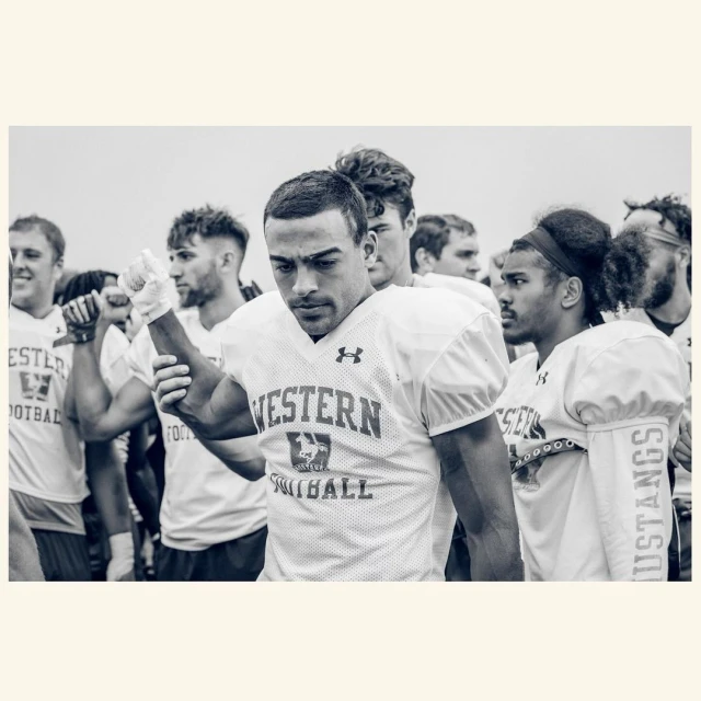 Black and white shot of Western Mustangs football players in uniform, focus on athlete in the foreground, sky background.