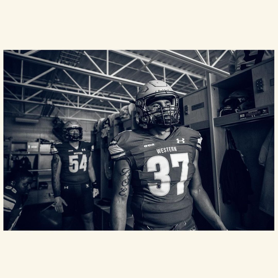 Black and white photo of two American football players in uniform in a locker room with metal lockers and overhead lighting.