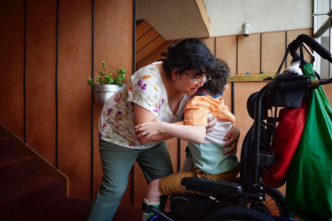Woman in floral top embraces child in wheelchair indoors, with plants and warm wood wall, creating a tender, supportive scene.
