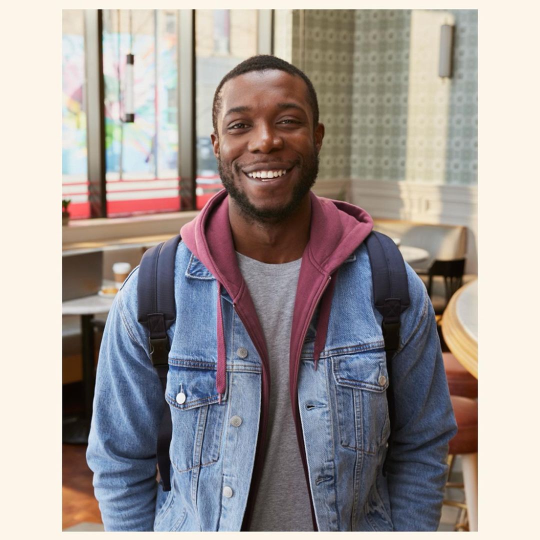 Smiling black man wearing a denim jacket and purple hoodie with a backpack inside a cafe in New York.