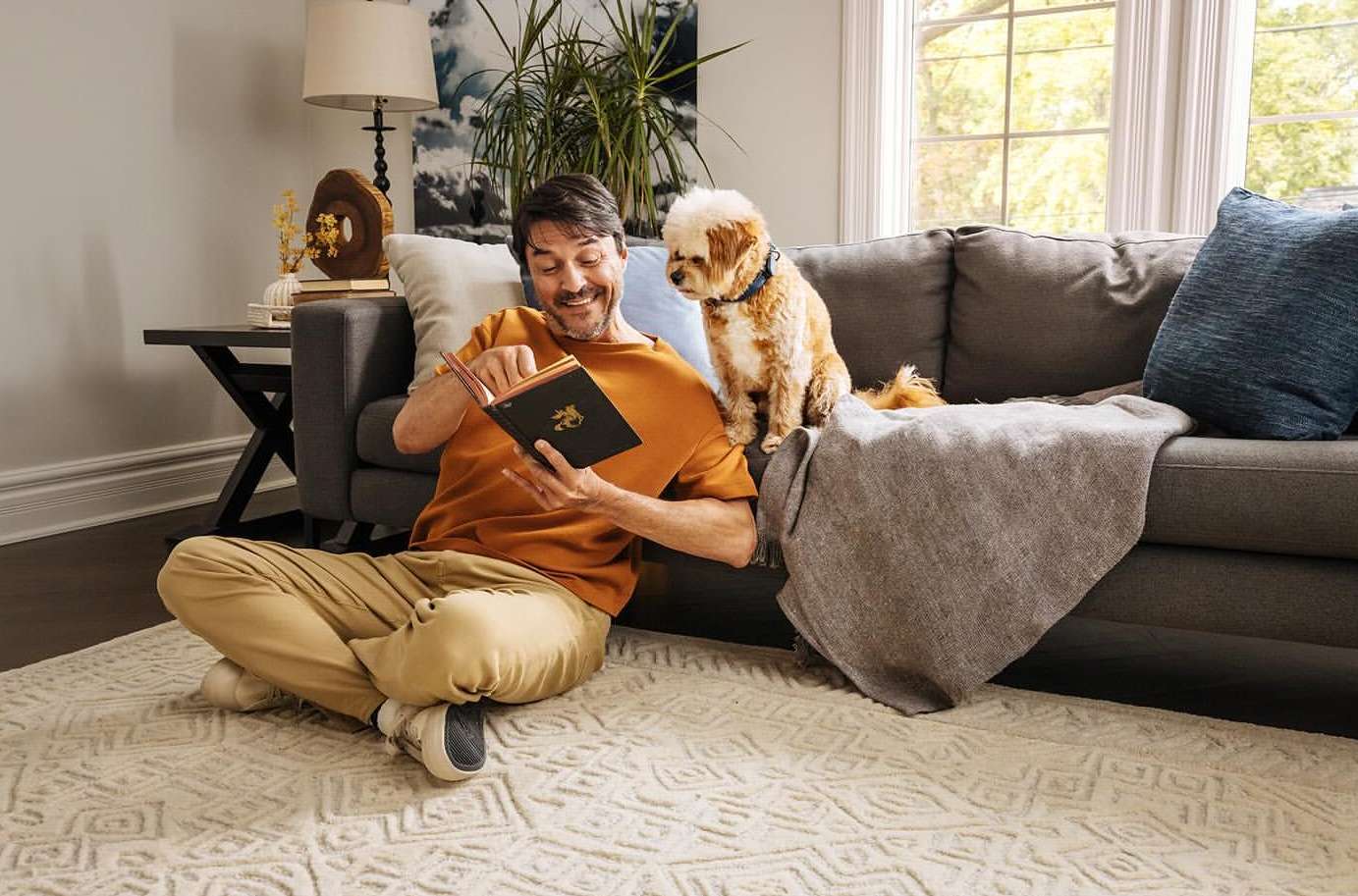 Man sits cross-legged on a patterned rug reading a book, with a small dog seated nearby on a gray sofa in a naturally lit living room.