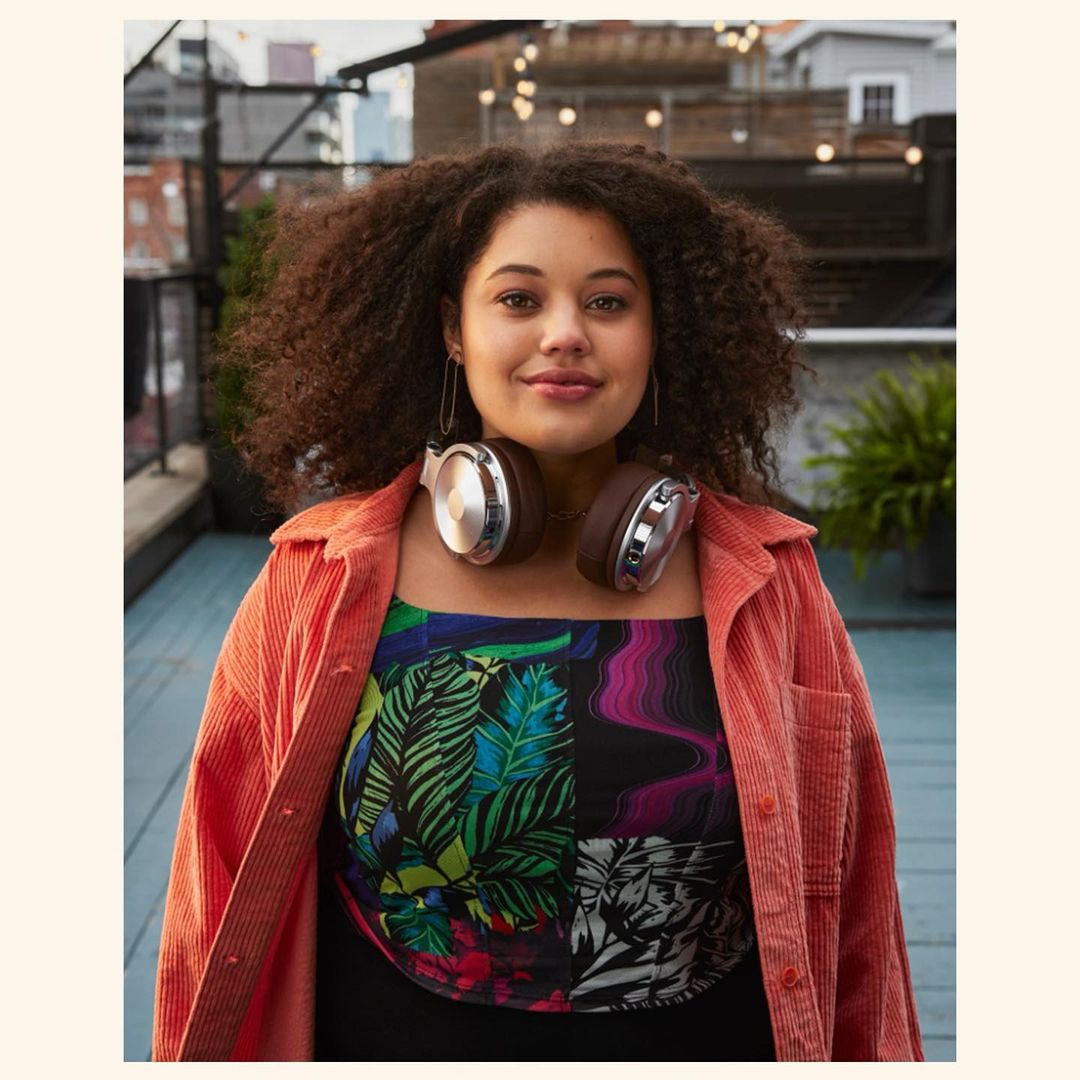 Woman with curly hair smiles wearing headphones, a colorful top, and a coral jacket on an urban rooftop with city buildings.