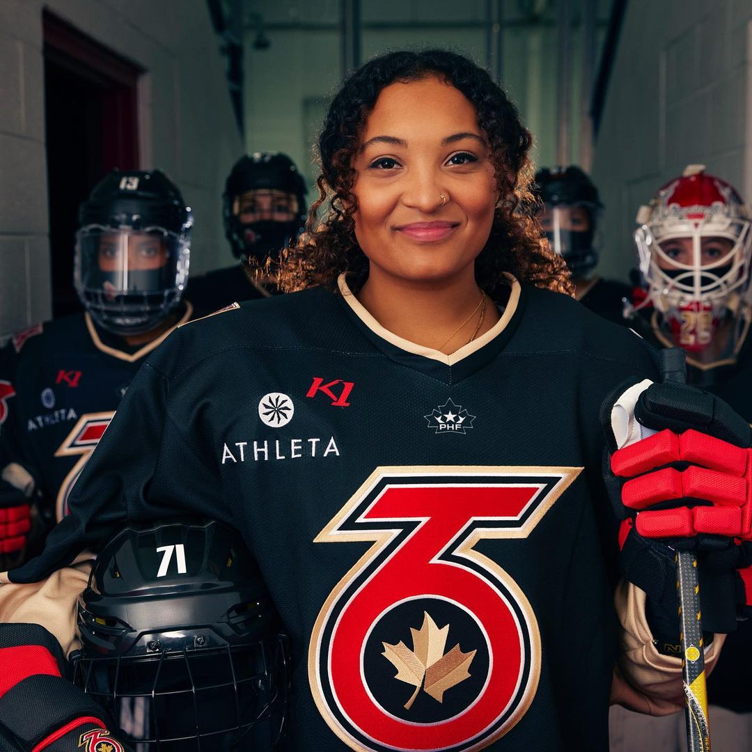 Portrait of a smiling hockey player in uniform in front of her team in a locker room, wearing a black, red, and gold jersey.