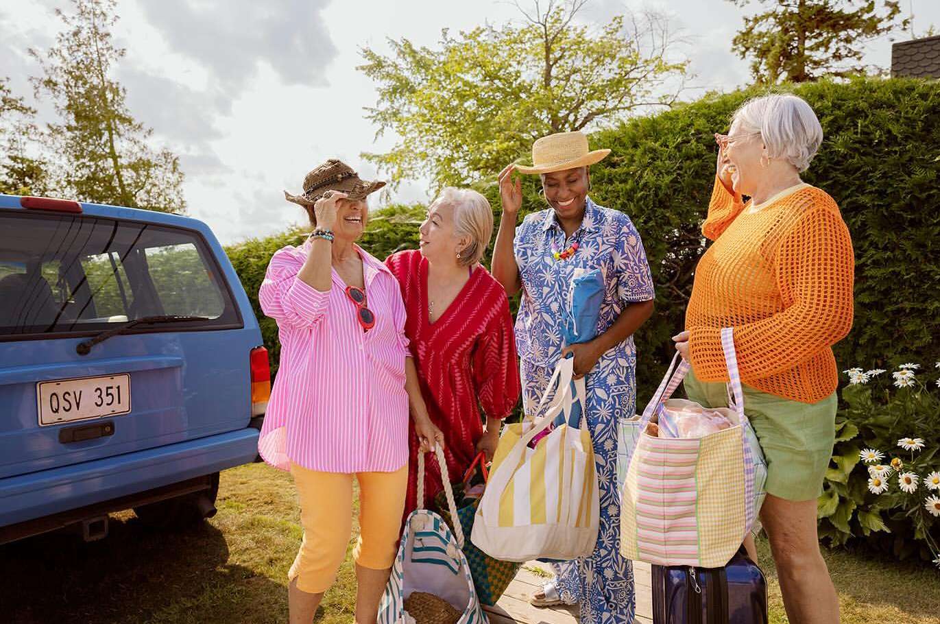 Four senior women in colorful vacation outfits unloading a blue SUV, standing outside on a sunny day with greenery in the background.