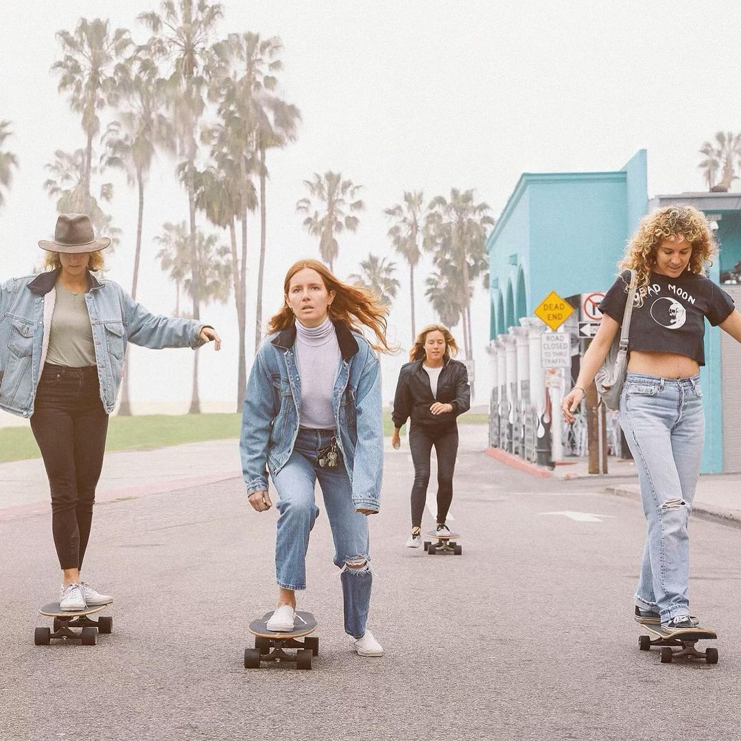 Four women skateboarding on a road near palm trees, wearing casual denim jackets and jeans with a building in the background.