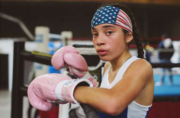 Young female boxer wearing pink boxing gloves and a US flag headband in a boxing ring, looking determined and ready for a fight.