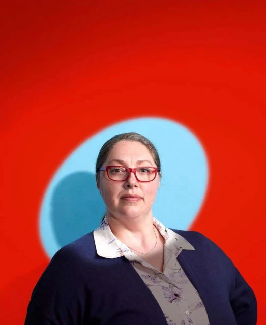 Portrait of an adult woman with red glasses in a floral shirt and blue cardigan, against a colorful red and blue backdrop in a studio.