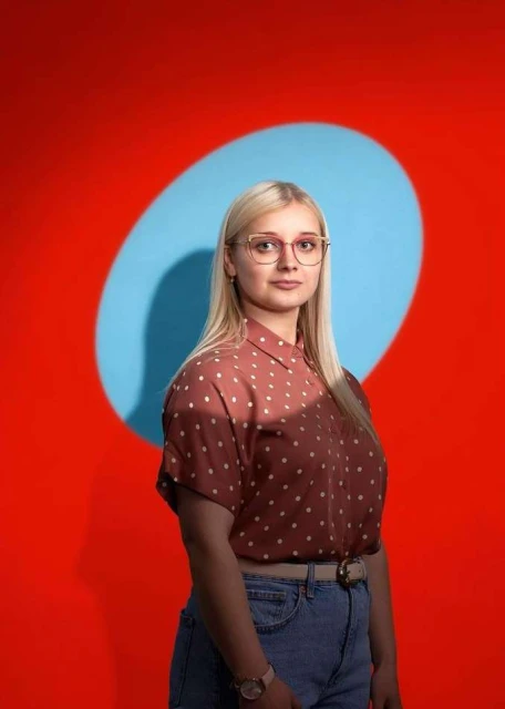 Portrait of a young woman with blonde hair and glasses, wearing a brown polka dot shirt, posing in front of a red background with a blue spotlight.