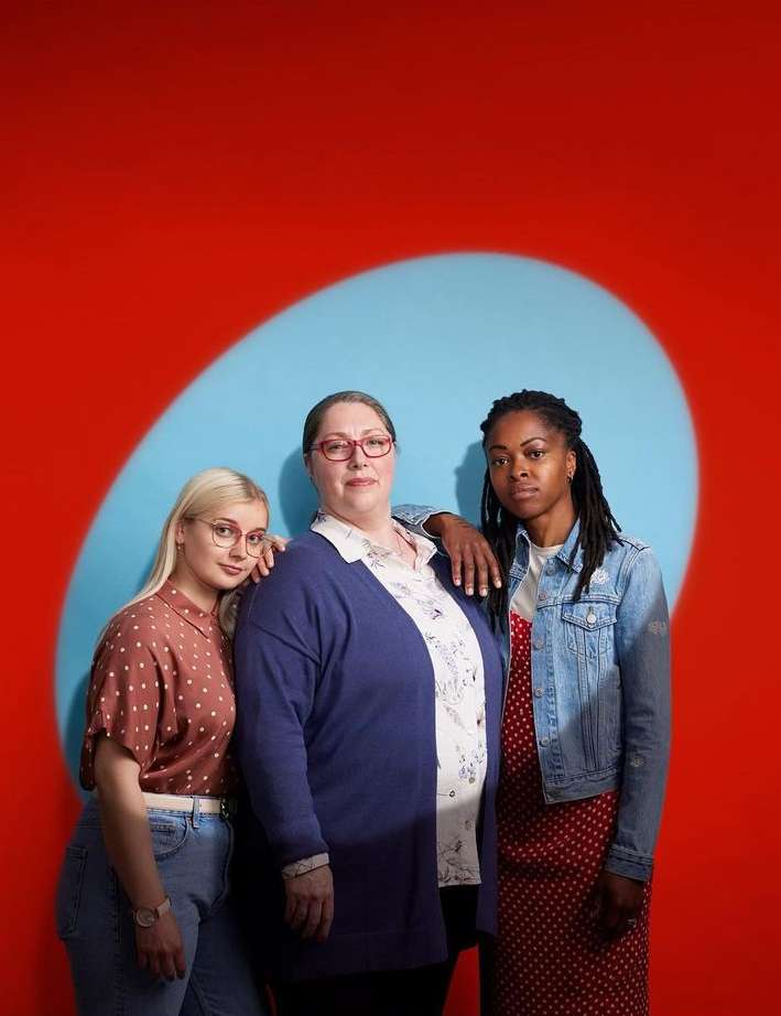 Three women stand in a studio setting. The woman in the center wears glasses and a cardigan. The background is red with a blue oval shape.