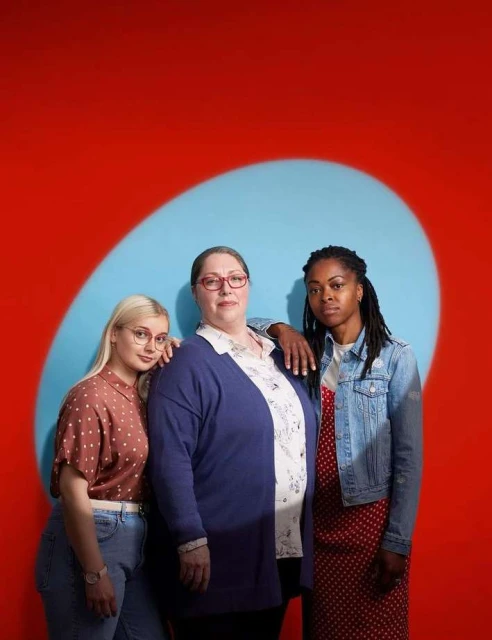 Three women stand in a studio setting. The woman in the center wears glasses and a cardigan. The background is red with a blue oval shape.