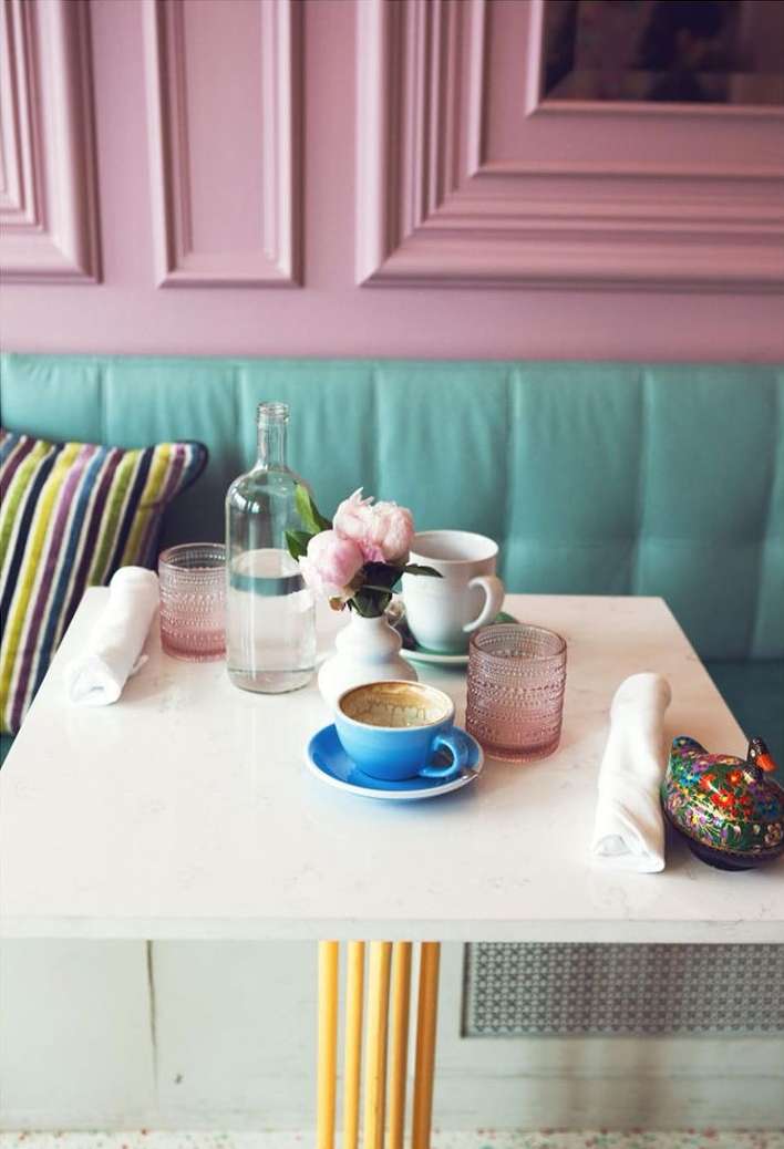 Cafe table setting with floral centerpiece, water bottle, glasses, and tableware in a pastel-colored interior. Soft, diffused light.