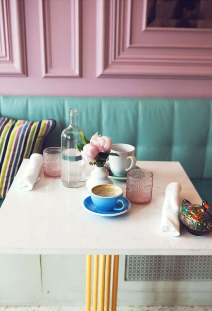 Cafe table setting with floral centerpiece, water bottle, glasses, and tableware in a pastel-colored interior. Soft, diffused light.