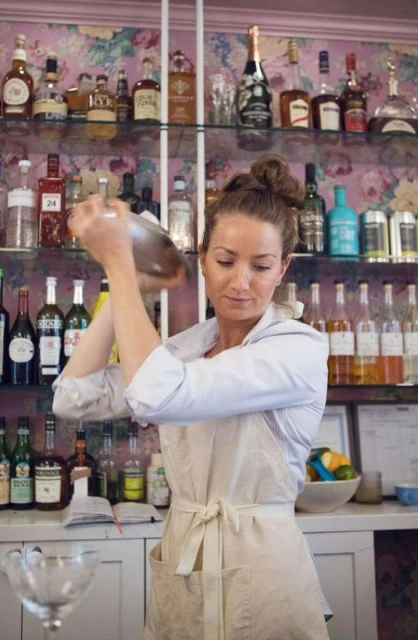 Woman bartender mixes drinks in a shaker in a bar with shelves of liquor bottles and floral wallpaper in soft, diffused light.