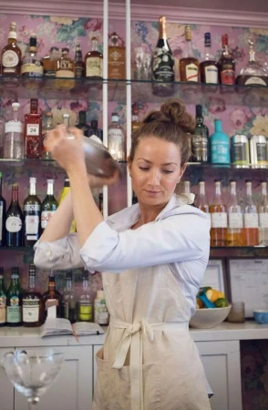 Woman bartender mixes drinks in a shaker in a bar with shelves of liquor bottles and floral wallpaper in soft, diffused light.