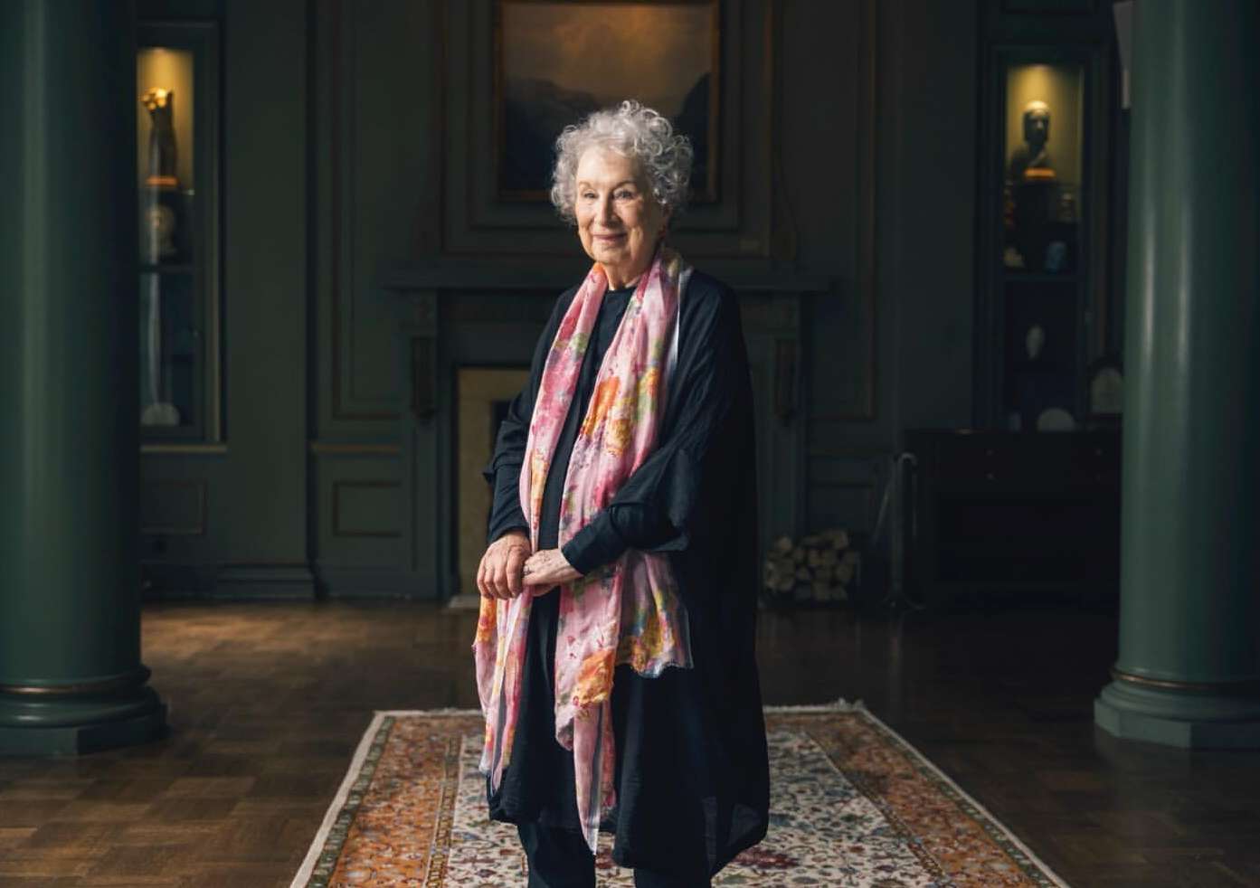 Elderly woman in a black dress and floral scarf stands in a grand interior with dark walls and columns, a fireplace, and antique furnishings.