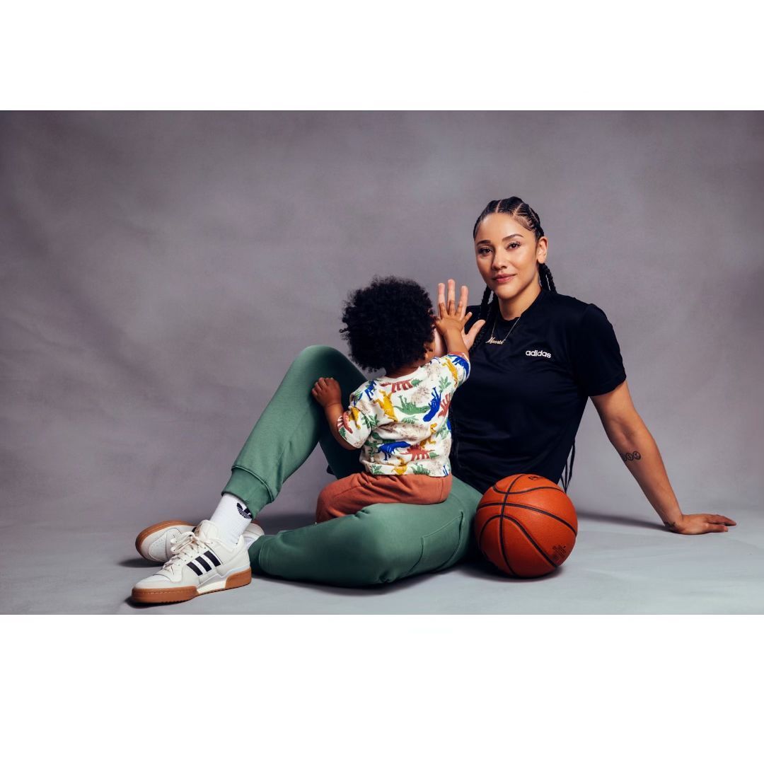 Adult woman sits with child on her lap in front of a grey backdrop. She wears athletic clothing. A basketball sits beside her.