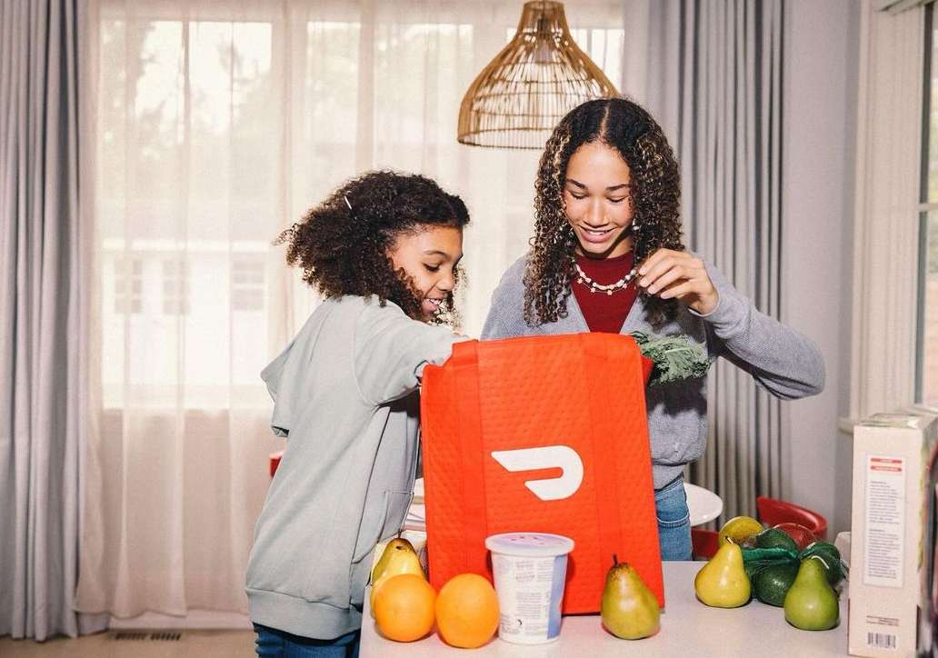 Two young girls open a DoorDash delivery bag at a kitchen counter with oranges, pears, and yogurt, with natural light from a window.