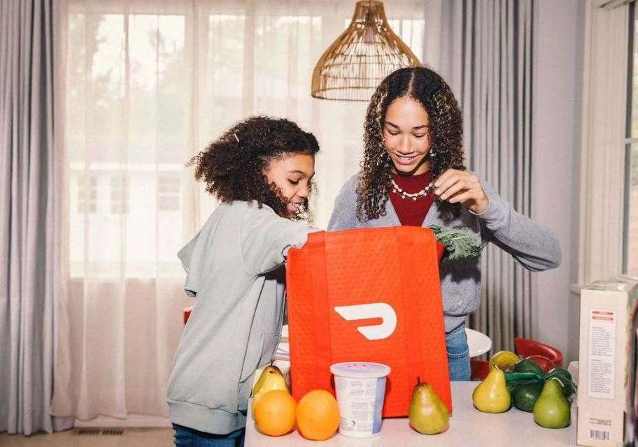 Two young girls open a DoorDash delivery bag at a kitchen counter with oranges, pears, and yogurt, with natural light from a window.