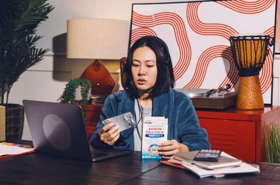 Asian woman in robe and headphones at desk with laptop and medicine, looking unwell in an indoor home setting.
