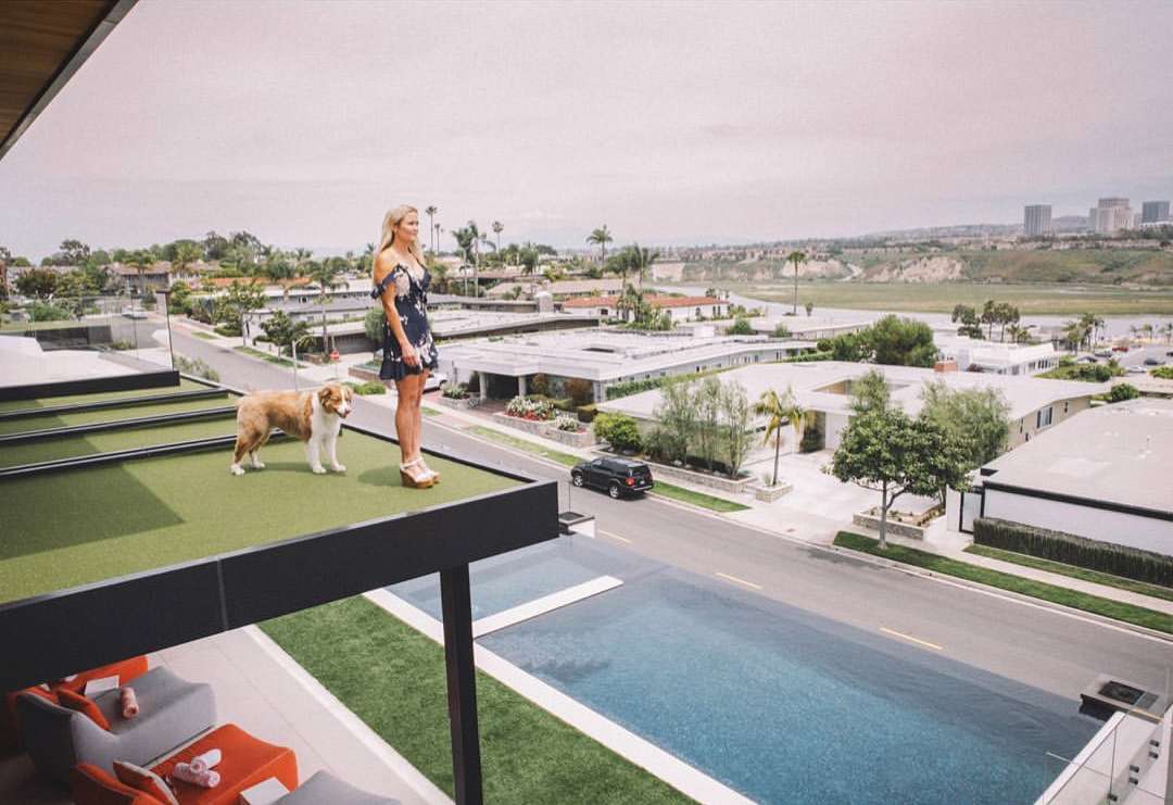 Woman and dog stand on modern rooftop overlooking a neighborhood and ocean, with a pool and lounge area visible below, in daytime.