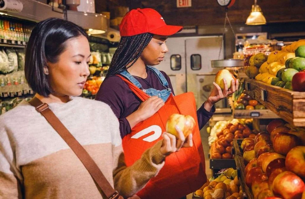 Two women shop for fruit at a grocery store. One holds an apple while wearing a red cap and a red delivery bag.