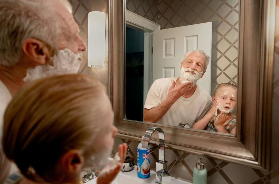Grandfather and grandson in bathroom, applying shaving cream to their faces, looking in the mirror in an indoor bathroom setting.