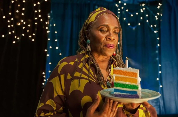 Older African-American woman holding rainbow cake with candle against background of blue drapes and string lights in an indoor environment.