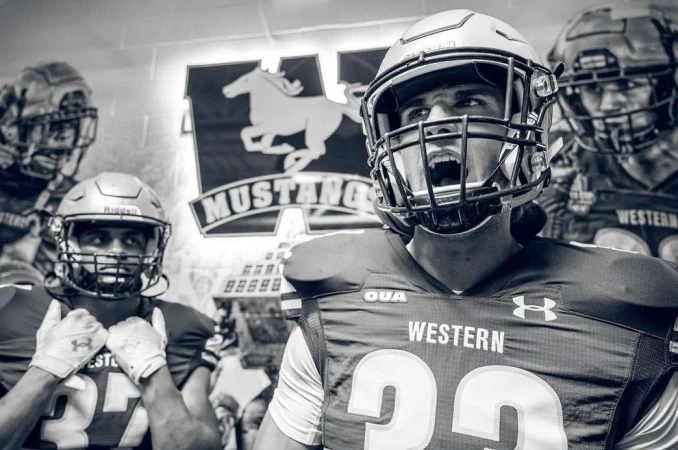 Monochrome shot of American football players with protective gear and intense facial expressions, posing with team logo in the background.