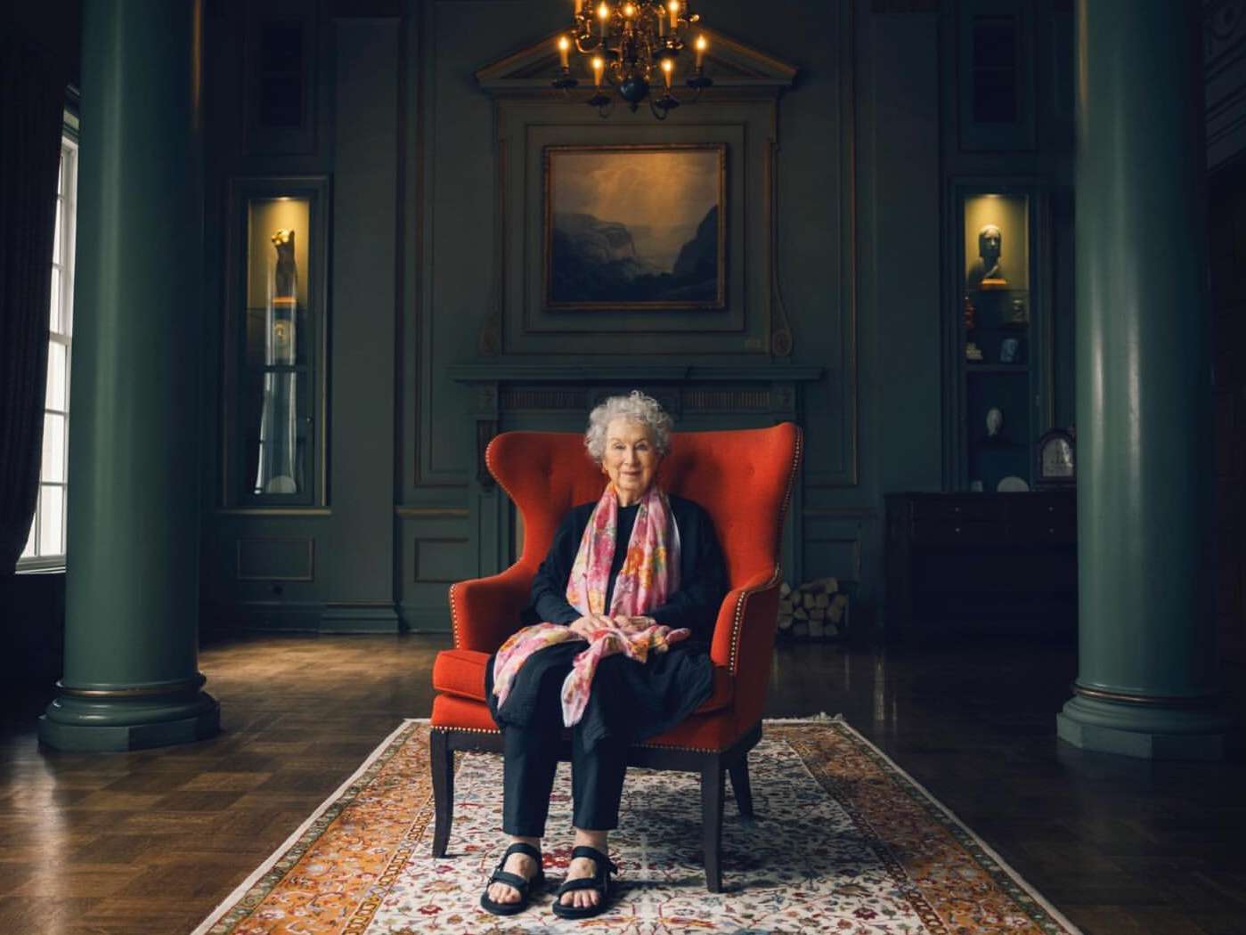 Elderly woman in a red armchair in a room with paneled walls, columns, hardwood floors, and a patterned rug.