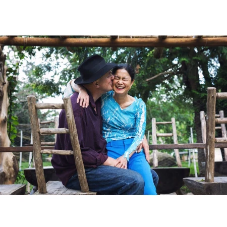Man wearing a black fedora kissing a laughing woman seated on his lap outdoors near rustic wooden railings.