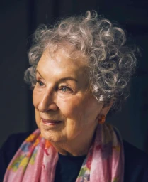 Portrait of an elderly woman with curly grey hair wearing a pink patterned scarf and looking thoughtfully to the side.