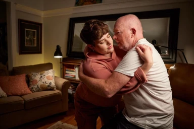 A person in a pink hoodie embraces an older man in a white t-shirt inside a softly lit living room.