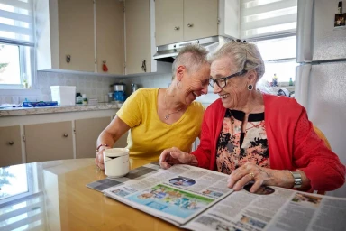 Two women laughing together at a kitchen table while reading a newspaper, photographed by Jennifer Roberts.