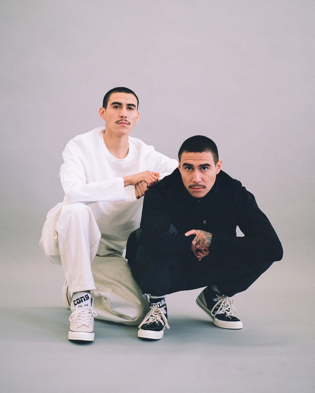 Two young men squat together in a studio setting, one in white, one in black, both sporting mustaches and wearing Converse sneakers.